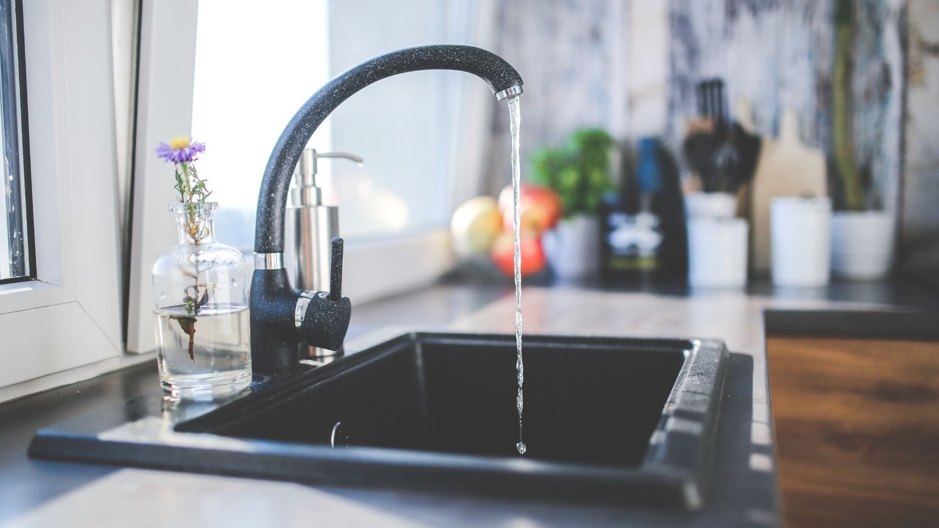 Black modern kitchen faucet dispensing water above dark sink near window.