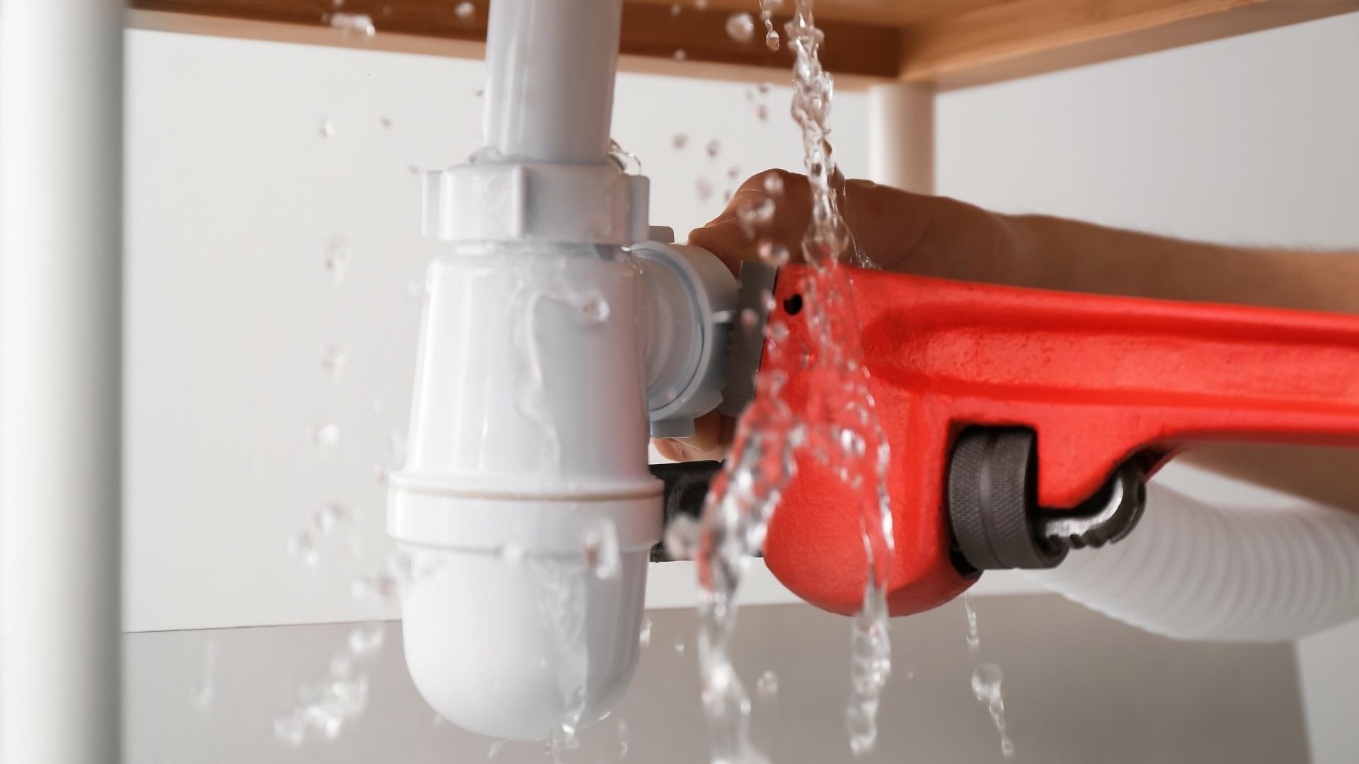 Person using red pipe wrench on white plumbing pipe with water flowing
